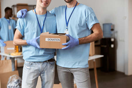 Cropped shot of two caucasian guys, male volunteers in blue uniform, protective gloves posing together and holding cardboard box for Donationの写真素材