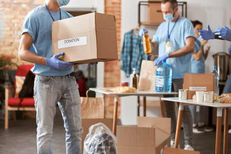 Cropped shot of male volunteer in blue uniform, protective mask and gloves holding cardboard box with donated food stuffs while standing indoorsの写真素材