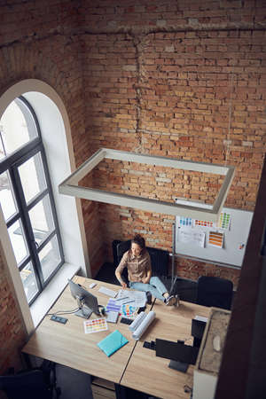 High angle view of relaxed woman designer taking a break while working on interior design project, sitting at the table in modern officeの写真素材