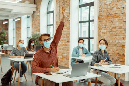 Male student in facial mask raising hand for answerの写真素材