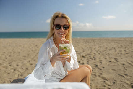 Portrait of lady drinking cocktail and smiling at camera while sunbathing on empty beachの写真素材