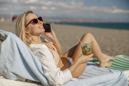 Smiling lady lying on deckchair, having call and drinking cocktail on a beachの写真素材