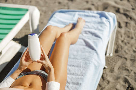 Woman in bikini holding sunscreen body lotion while lying on deck chair