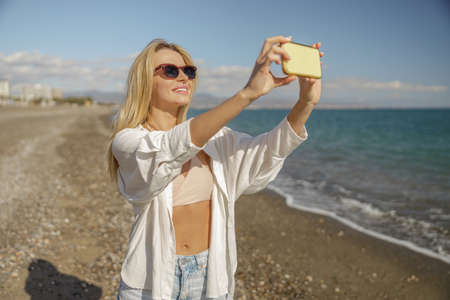 Cheerful female tourist taking pictures of the sea coast using smartphone, standing on the beachの写真素材