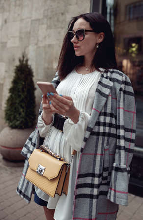 Beautiful stylish female in sunglasses texting on smartphone at street. City life.
