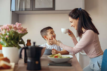 Beautiful mother drinking coffee in the morning and cleaning cheek of small son.の写真素材