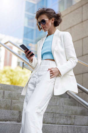 Attractive businesswoman in white suit using phone standing near business centreの写真素材