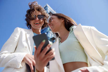 Smiling female friends in trendy outfits use phone while relaxing in a city