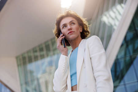 Attractive young businesswoman in white suit talking phone standing near business centreの写真素材