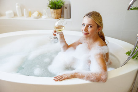 Attractive young blonde female sitting in bathtub with foam and holding white wine glass.の写真素材