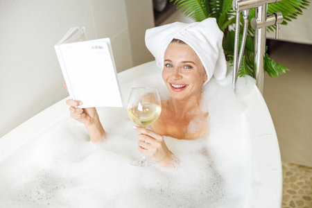 Portrait of pretty smiled young female lying in bath with foam, reading and drinking wine.の写真素材