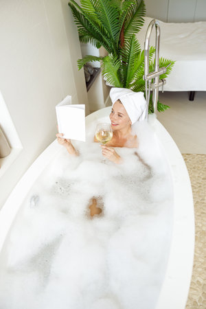 Attractive Caucasian young woman in towel on head taking bath and reading book with wine.の写真素材