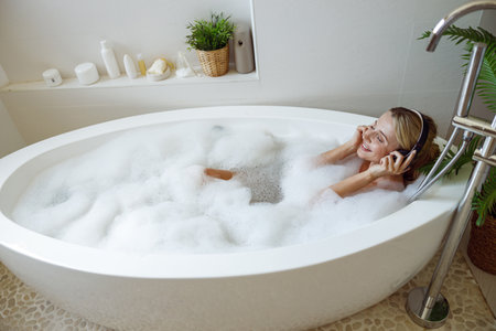 Caucasian young pretty woman in headphones lying in bathtub with foam and listening to musicの写真素材