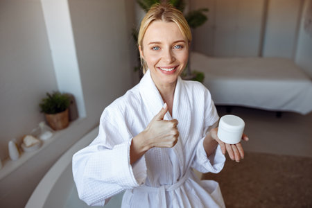 Cheerful young Caucasian woman smiling, holding cream and giving the thumb up. Spa.の写真素材