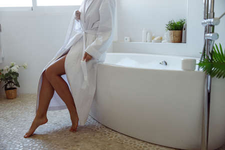 Caucasian female in white gown sitting at bathtub with bubbles and foam. Naked legs concept.