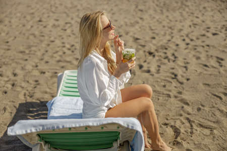 Blonde lady enjoying mohito cocktail and looking away on empty beachの写真素材