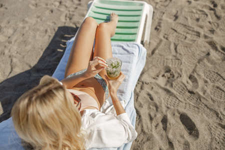 Top view of tanned young woman in bikini with drink sitting on a deck chair, enjoying sunbath