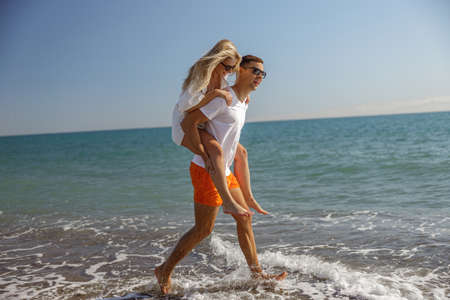 Smiling man in sunglasses carrying his woman on his back while walking together along sea coastの写真素材