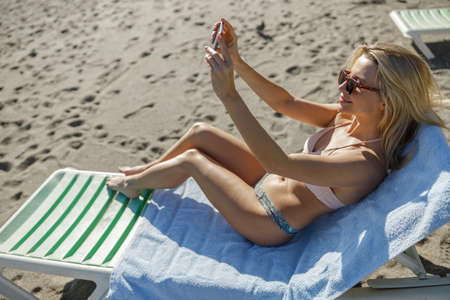 Cheerful woman on vacation taking selfie photos using smartphone while relaxing on the beachの写真素材