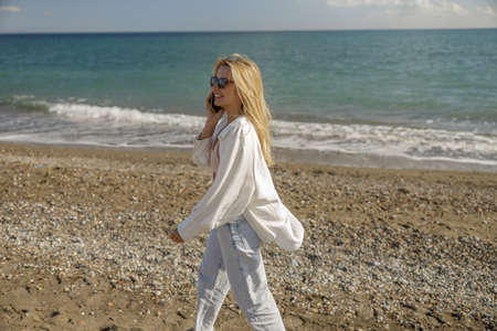 Content lady in sunglasses having a call, using smartphone while walking along the beach aloneの写真素材