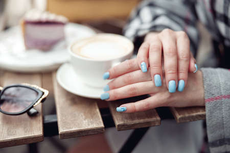 Close up of female hands with blue manicure and cup of coffee at cafe. Cappuccino time.の写真素材