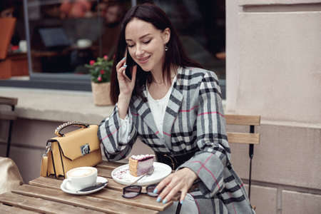 Smiled young pretty woman having phone talk while eating cheesecake with cappuccino in cafe.の写真素材