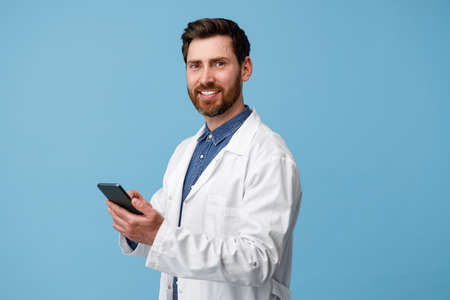 Cheerful man doctor holding mobile phone over blue studio backgroundの写真素材