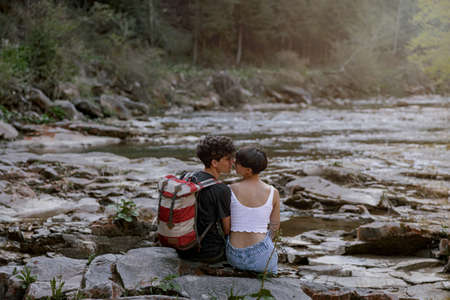 Back view of couple of hikers sitting on bank of rocky river and kissing. Hiking in mountains.の写真素材