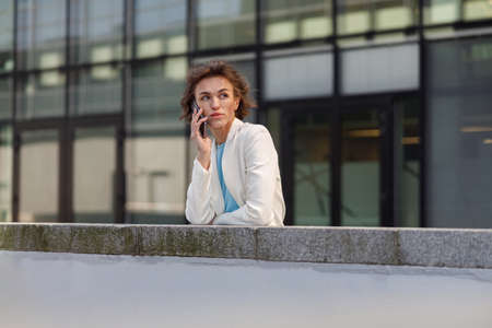 Attractive young businesswoman in white suit talking phone standing near business centre