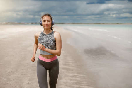 Young beautiful sportive woman in headphones running over seaside on morning at beach