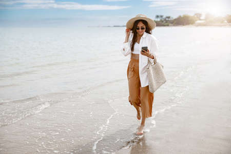 Cheerful young woman traveler with straw hat walking on beach during vacation and looking phoneの写真素材