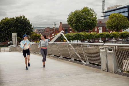 Two athletic women in sportswear is jogging on a bridge and listening music. Reaching the goal