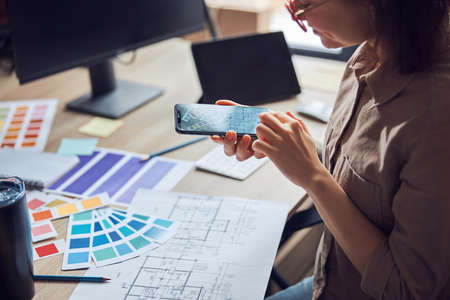 Woman taking a photo of a blueprint and color samples palette using smartphone while working on new interior design project in her officeの写真素材