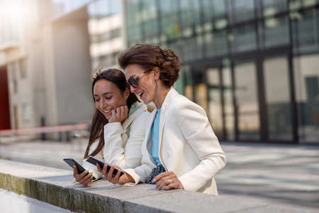Smiling female friends in trendy outfits use their phones while relaxing in a cityの写真素材