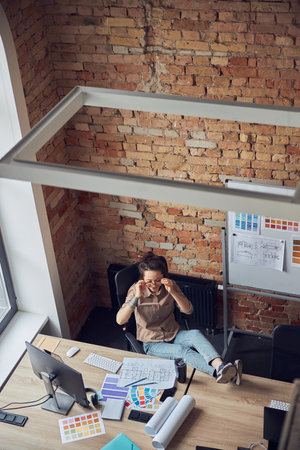 High angle view of creative female designer putting on glasses while working on interior design project, sitting at the table in modern officeの写真素材