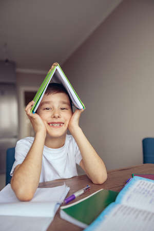 Portrait of cute small Caucasian boy with braces holding textbook over head and smiling.