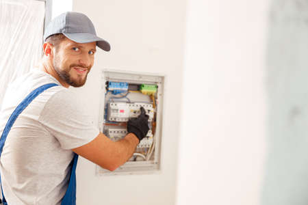 Smiling electrician looking at camera while working with fuses at the circuit breaker control cabinet on the wallの写真素材