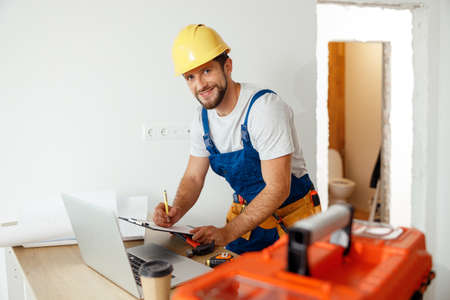 Smiling handyman worker in uniform and hard hat using laptop, writing on a clipboard, looking at...