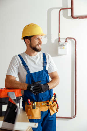 Smiling technician, handyman in uniform, hard hat and protective gloves looking away while standing indoors, leaning on a toolboxの写真素材