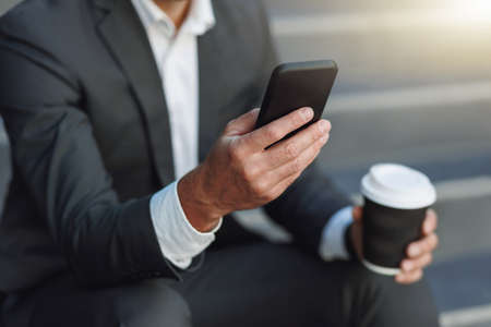 Close up of office worker in suit sitting on stairs with coffee during break and using phoneの写真素材