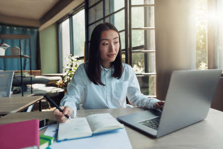 Attractive asian businesswoman working on laptop and making notes at modern office