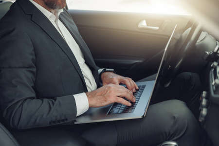 Close up of businessman working on laptop while sitting on driver seat in luxury carの写真素材