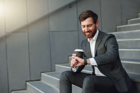 Attractive office worker in suit sitting on stairs with coffee during break and looking on his watchの写真素材