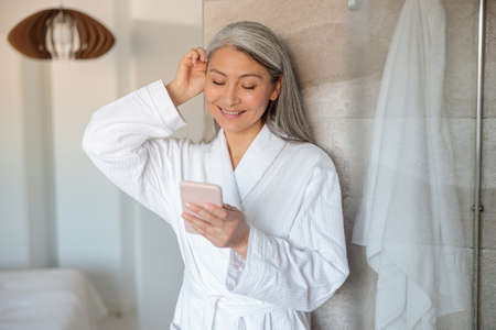 Pretty Asian woman in bathrobe halding smartphone in showerの写真素材