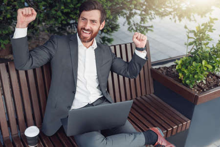 Businessman rejoices over completion of work on project while sitting on bench near officeの写真素材