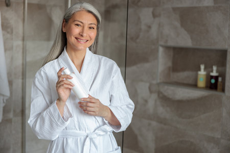 Smiling Asian lady standing in bathroom with shampooの写真素材