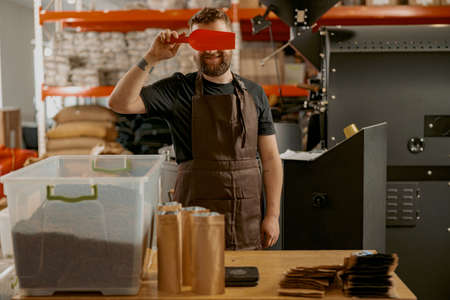 Playful barista closes eyes with a small ladle while standing at a small coffee factory