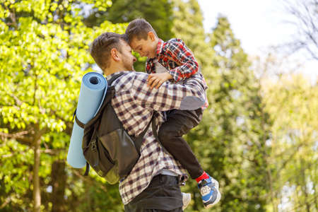 Cheerful Caucasian father with backpack holding small son on hands in park on summer day.の写真素材