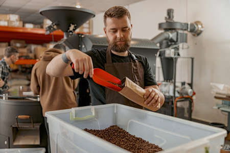 Barista packs roasted coffee beans into packages for sale on background of roasting machine