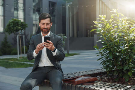 Smiling office worker have a rest during break outside office and scrolling social media on phoneの写真素材